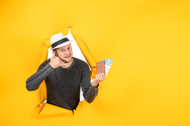 Widok z przodu uśmiechniętego młodego mężczyzny w kapeluszu trzymającego zagraniczny paszport z biletem i wykonującego gest „Zadzwoń do mnie” w rozdartej na żółtej ścianie