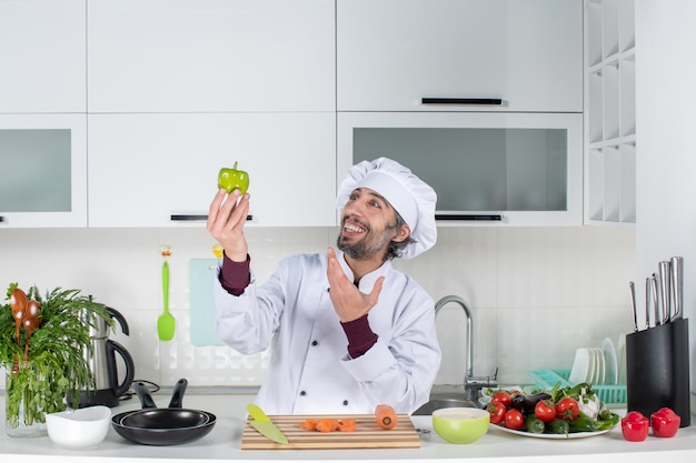 Widok z przodu uśmiechnięty kucharz w mundurze trzymający zielony pieprz w kuchni