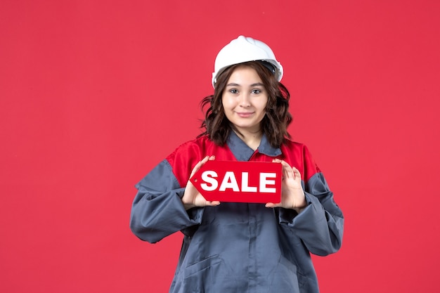 Widok z przodu z bliska pewna pracownica w mundurze w kasku pokazująca ikonę sprzedaży na odizolowanej czerwonej ścianie