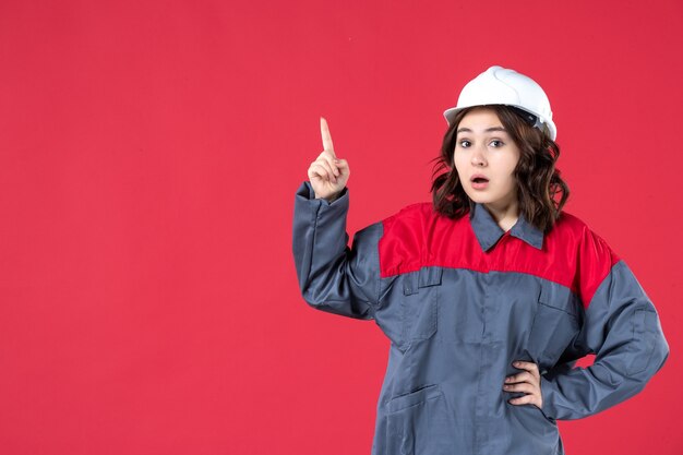 Widok z przodu zaskoczonej kobiety budowniczej w mundurze z twardym kapeluszem i wskazującej na pojedyncze czerwone tło