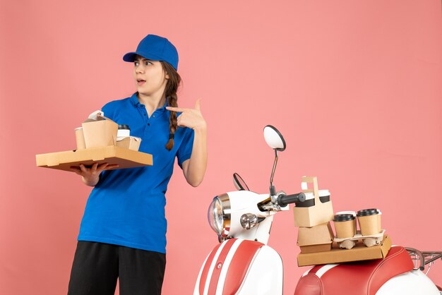 Widok z przodu zastanawiającej się kurierki stojącej obok motocykla trzymającego kawę i małe ciastka na pastelowym brzoskwiniowym tle