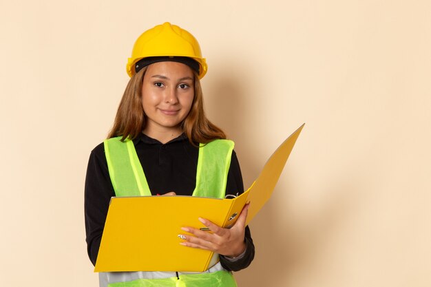 Widok z przodu żeński konstruktor w żółtym kasku trzymający żółty plik zapisujący notatki uśmiechnięte na białej ścianie