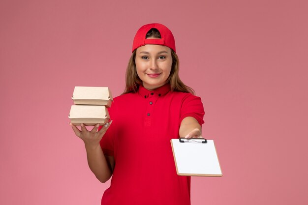 Bezpłatne zdjęcie widok z przodu żeński kurier w czerwonym mundurze i pelerynie trzymający małą dostawę paczek z żywnością notatnik na jasnoróżowej ścianie, jednolita usługa dostarczania pracy pracownika