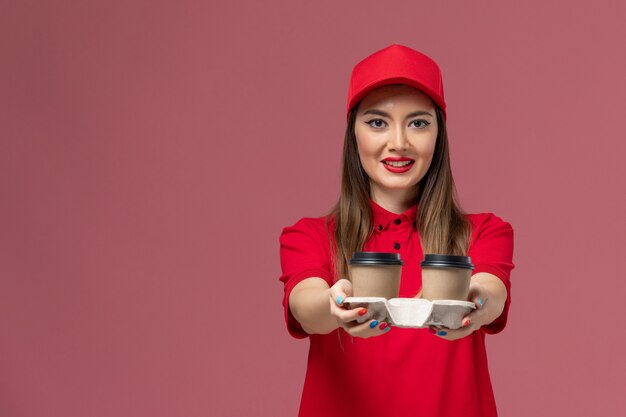 Widok z przodu żeński kurier w czerwonym mundurze, trzymając filiżanki kawy dostawy z uśmiechem na różowym tle, pracownik służbowy mundur dostawy