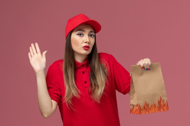 Widok z przodu żeński kurier w czerwonym mundurze, trzymając papierowy pakiet żywności na jasnoróżowym tle, pracownik usługi dostawy pracy firma mundurowa