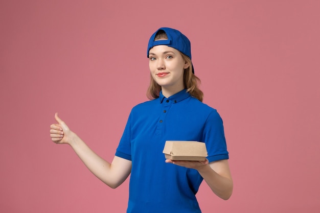 Widok z przodu żeński kurier w niebieskim mundurze i pelerynie trzyma mały pakiet żywności dostawy na różowym tle jednolite usługi dostawy pracy firmy