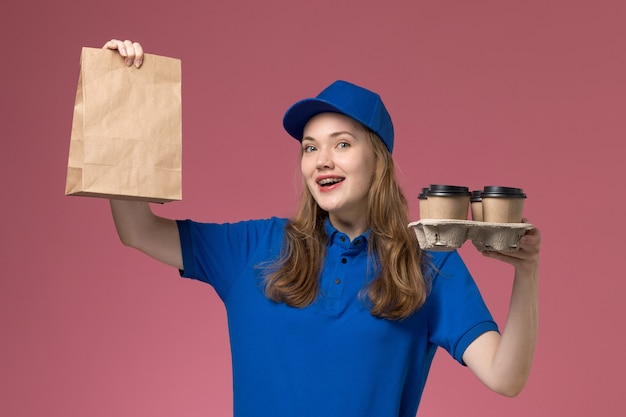 Bezpłatne zdjęcie widok z przodu żeński kurier w niebieskim mundurze trzyma brązowe kubki z dostawą kawy z pakietem żywności i uśmiech na różowym biurku jednolity pracownik firmy