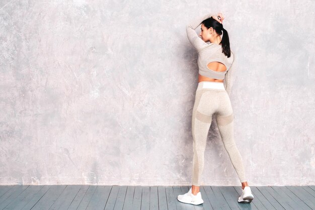 Widok z tyłu fitness pewnie kobieta w sportowej odzieży Sexy młody piękny model z idealnym ciałem Kobieta pozuje w pobliżu szarej ściany w studio Rozciąganie się przed treningiem