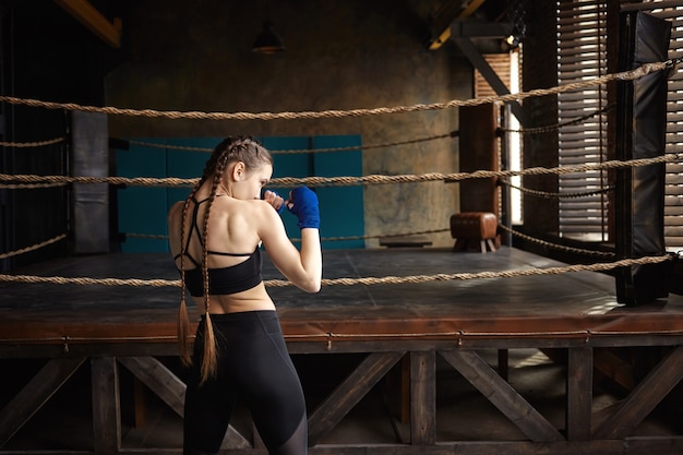 Widok z tyłu zawodowej bokserki z dwoma warkoczami stojącej w pustej siłowni z ringiem w tle, sam trenujący