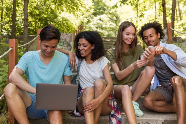 Wieloetniczni przyjaciele uczniowie na zewnątrz za pomocą telefonu komórkowego i laptopa