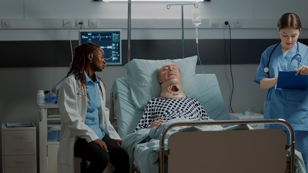 Bezpłatne zdjęcie wieloetniczny personel medyczny sprawdzający opiekę zdrowotną pacjenta na szpitalnym łóżku na oddziale intensywnej terapii. afroamerykański lekarz pomaga starcowi z kołnierzem szyjnym i rurką tlenową
