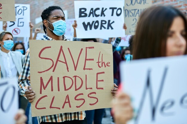 Wieloetniczny tłum ludzi protestujących przeciwko bezrobociu z powodu pandemii koronawirusa W centrum uwagi jest czarnoskóry mężczyzna trzymający transparent z napisem „ratuj klasę średnią”
