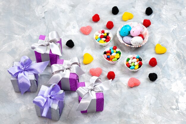 Wielokolorowe cukierki z widokiem z góry na małych talerzach wraz z marmoladami w kształcie serca i fioletowymi pudełkami na prezenty na szarym tle