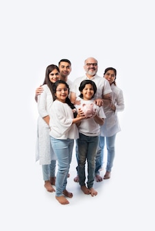 Wielopokoleniowa indyjska sześcioosobowa rodzina trzymająca skarbonkę w białych ubraniach i stojąca przy białej ścianie