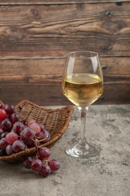 Wiklinowy kosz czerwonych winogron z lampką białego wina na marmurowym stole.