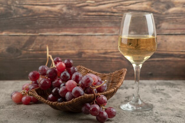 Wiklinowy kosz czerwonych winogron z lampką wina na marmurowym stole.