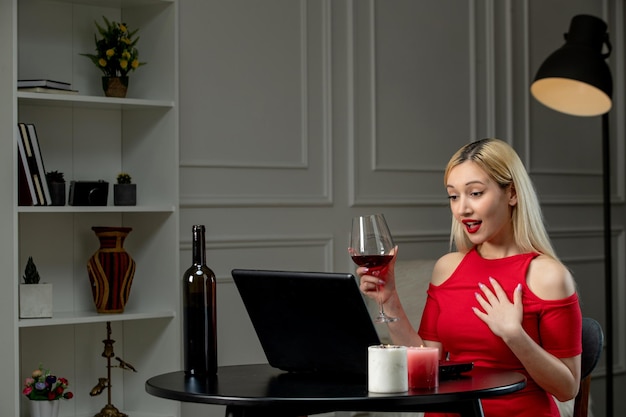 Bezpłatne zdjęcie wirtualna miłość ładna blondynka w czerwonej sukience na randkę na odległość z winem wskazującym na siebie
