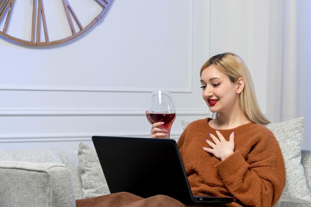 Wirtualna miłość ładna młoda blondynka w przytulnym swetrze na komputerowej dacie odległości trzymającej kieliszek do wina