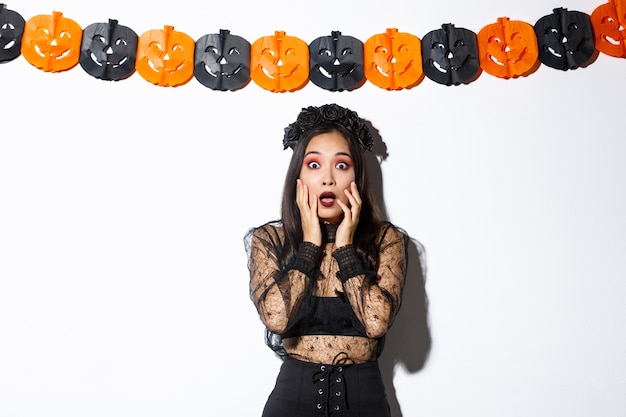 Wizerunek kobiety w stroju czarownicy wyglądającej na przestraszoną, wyrażającej przerażenie lub strach, stojąc na białym tle z dekoracją banerów dyni, świętując Halloween.