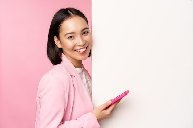Wizerunek koreańskiej kobiety przedsiębiorcy w garniturze stojącej w pobliżu reklamy ściennej na tablicy trzymającej smartfona i uśmiechającej się pozowanie na różowym tle
