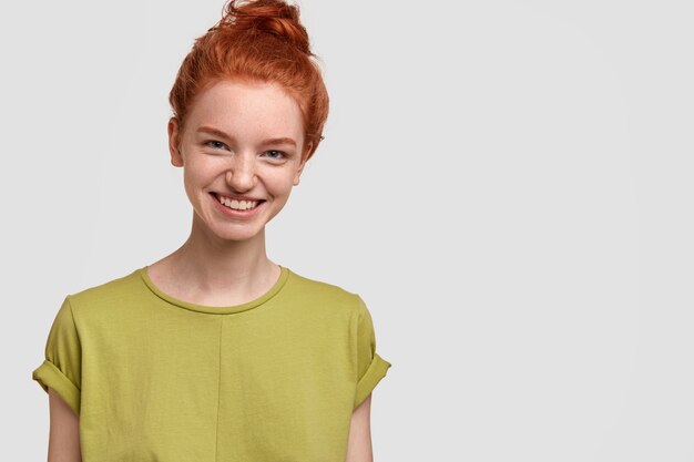 Wizerunek ładnej rudowłosej dziewczyny ma czarujący uśmiech na twarzy, nosi zwykłą zieloną koszulkę, czuje się szczęśliwa, odizolowana na białej ścianie z wolnym miejscem na treść reklamową lub promocję. Emocje