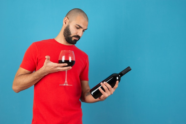 Wizerunek młodego mężczyzny modelu w czerwonym t-shorcie trzyma butelkę wina ze szkłem.