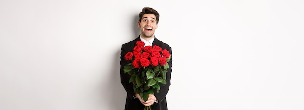 Bezpłatne zdjęcie wizerunek przystojnego chłopaka w czarnym garniturze trzymającego bukiet czerwonych róż i uśmiechającego się na randce