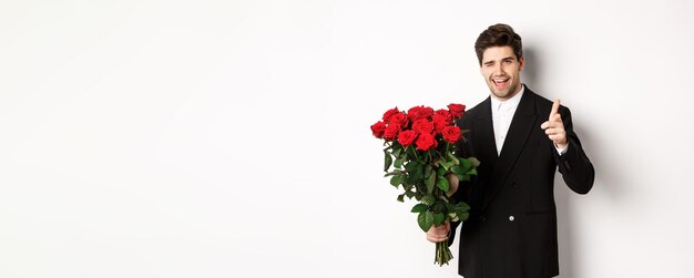 Wizerunek przystojnego romantycznego faceta w czarnym garniturze trzymającego bukiet róż i wskazującego na kamerę gratulacje