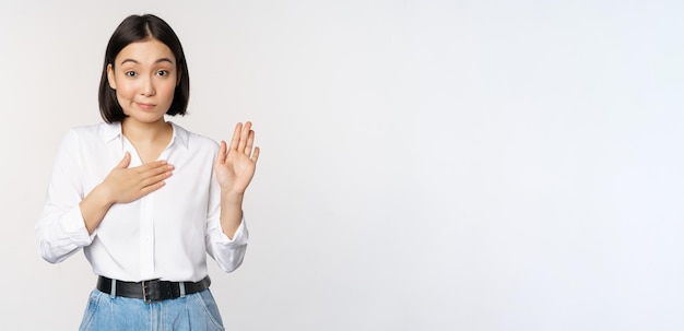 Wizerunek ślicznej młodej pracowniczki biurowej azjatyckiej studentki podnoszącej rękę do góry i kładącej dłoń na klatce piersiowej, która sama przedstawia składając obietnicę stojącą na białym tle