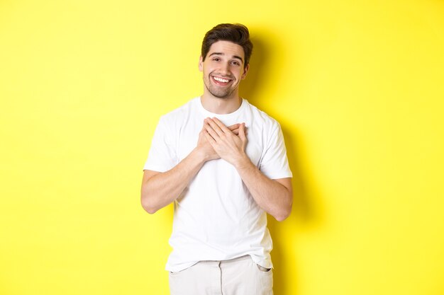 Wizerunek wdzięcznego przystojnego faceta w białej koszulce, trzymającego ręce na sercu i uśmiechniętego zadowolonego, wyrażającego wdzięczność, dziękującego za coś, stojącego na żółtym tle.