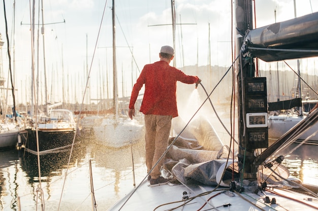 Właściciel jachtu lub żeglarz używa węża do mycia słonej wody z pokładu jachtu, gdy jest on zacumowany lub zaparkowany w marinie o zachodzie słońca