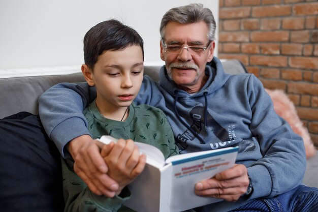Wnuk czytanie książki ze swoim szczęśliwym dziadkiem siedząc na kanapie.