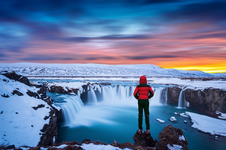 Bezpłatny plik Zdjęcie wodospad godafoss o zachodzie słońca w zimie, islandia. facet w czerwonej kurtce patrzy na wodospad godafoss.
