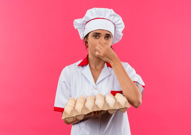 Wrażliwa młoda kobieta kucharz w mundurze szefa kuchni, trzymając partię jaj i zamknięty nos na izolowanej różowej ścianie z miejsca na kopię