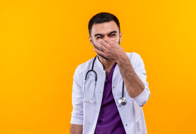 Wrażliwy młody mężczyzna lekarz ubrany w stetoskop medyczny fartuch zamknięty nos na na białym tle żółtym tle