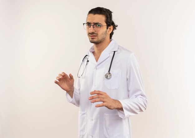 Wrażliwy młody mężczyzna lekarz z okularami optycznymi na sobie białą szatę ze stetoskopem na odosobnionej białej ścianie z miejsca na kopię