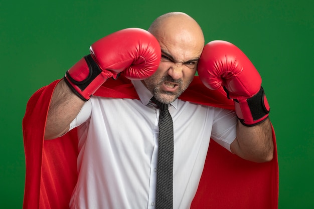 Bezpłatne zdjęcie wściekły biznesmen superbohatera w czerwonej pelerynie i rękawicach bokserskich uderzając się, robiąc grymas stojąc nad zieloną ścianą