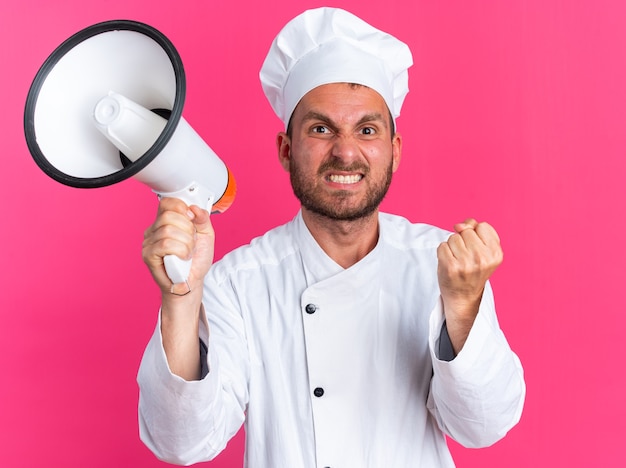 Wściekły młody kucharz rasy kaukaskiej w mundurze szefa kuchni i czapce, trzymający głośnik zaciskający pięść
