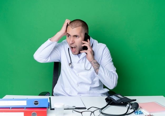 Bezpłatne zdjęcie wściekły młody lekarz płci męskiej ubrany w szlafrok medyczny i stetoskop siedzi przy biurku z narzędziami roboczymi rozmawia przez telefon kładąc rękę na głowie patrząc na bok odizolowany na zielono