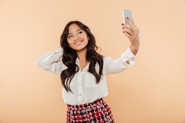 Wspaniała azjatykcia kobieta z długim ciemnym włosy robi selfie fotografii na jej smartphone ono podziwia nad beżowym tłem