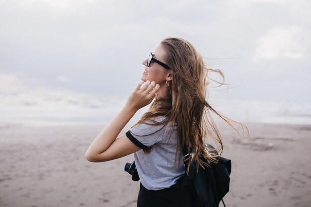 Wspaniała fotografka w szarym t-shircie patrząc na zachmurzone niebo. Zewnątrz portret romantycznej dziewczyny brunetka z aparatu, zabawy na plaży w zimny dzień.