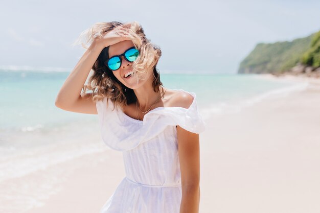Wspaniała kobieta w białej sukni dotyka jej włosów na morzu. Opalona beztroska kobieta w okularach przeciwsłonecznych wyrażająca radość z wakacji.