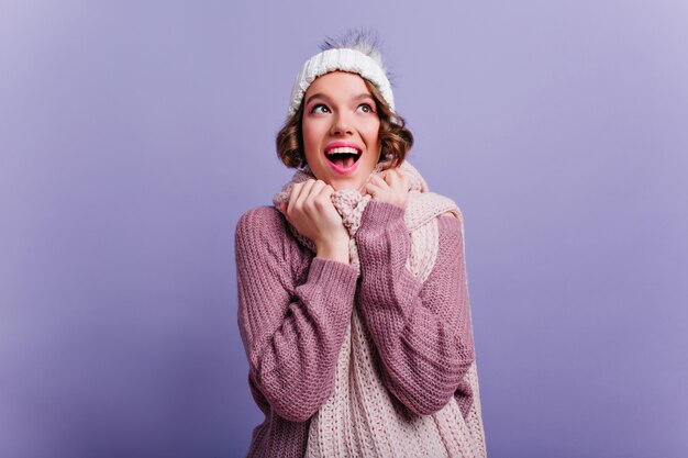 Wspaniała modelka wyrażająca radosne emocje podczas sesji zdjęciowej w zimowych ubraniach. Wewnątrz portret ładnej dziewczyny ze stylową fryzurą nosi miękki sweter.