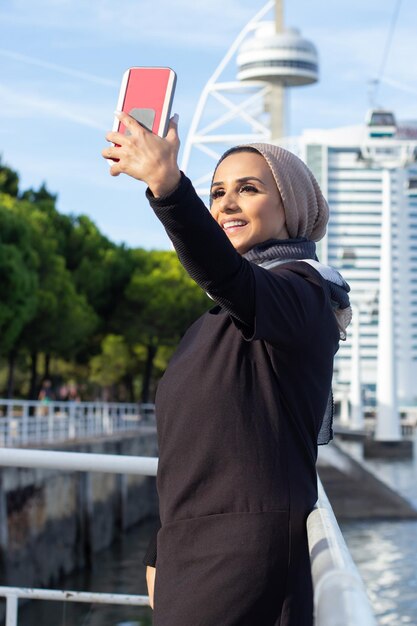 Wspaniała uśmiechnięta Arabka biorąc selfie. Kobieta z zakrytą głową i makijażem, wyobrażając sobie siebie z telefonem komórkowym. Międzynarodowa, piękna koncepcja mediów społecznościowych