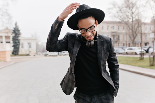 Wspaniały afrykański mężczyzna z nieśmiałym uśmiechem na ulicy miasta. Stylowy czarny facet w kapeluszu stojący na drodze ze słuchawkami i śmiejąc się.