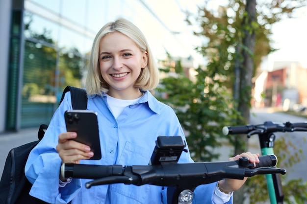 Bezpłatne zdjęcie współczesna młoda kobieta skanuje kod qr za pomocą aplikacji na smartfon, aby wynająć skuter elektryczny na ulicy, uśmiechając się
