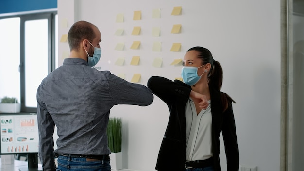 Współpracownicy z maseczką dotykają łokcia ze swoim kolegą, aby zapobiec zakażeniu koronawirusem. Współpracownicy szanujący dystans społeczny podczas pracy nad firmowym projektem komunikacyjnym