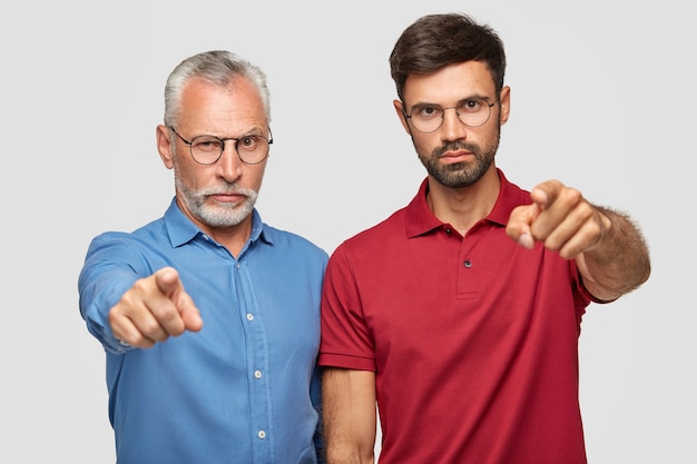 Wybieramy dokładnie Ciebie! Dwaj pewni siebie poważni mężczyźni wskazują palcami wskazującymi, wyrażają swój wybór, noszą jasne ubrania, odizolowani na białej ścianie. Starszy mężczyzna z synem dorosłych w pomieszczeniu