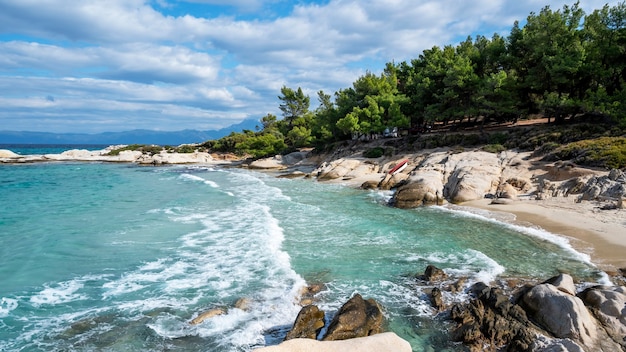 Wybrzeże Morza Egejskiego otoczone zielenią, skałami, krzewami i drzewami, błękitna woda z falami, Grecja