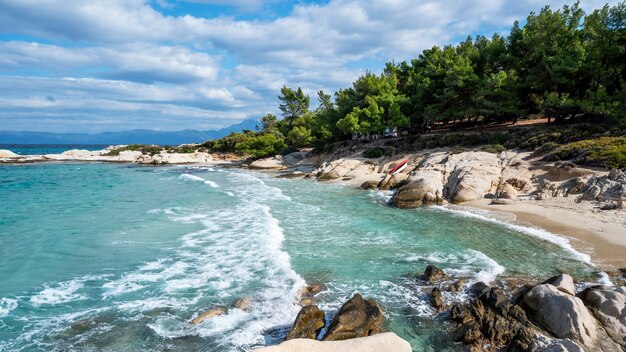 Wybrzeże Morza Egejskiego otoczone zielenią, skałami, krzewami i drzewami, błękitna woda z falami, Grecja